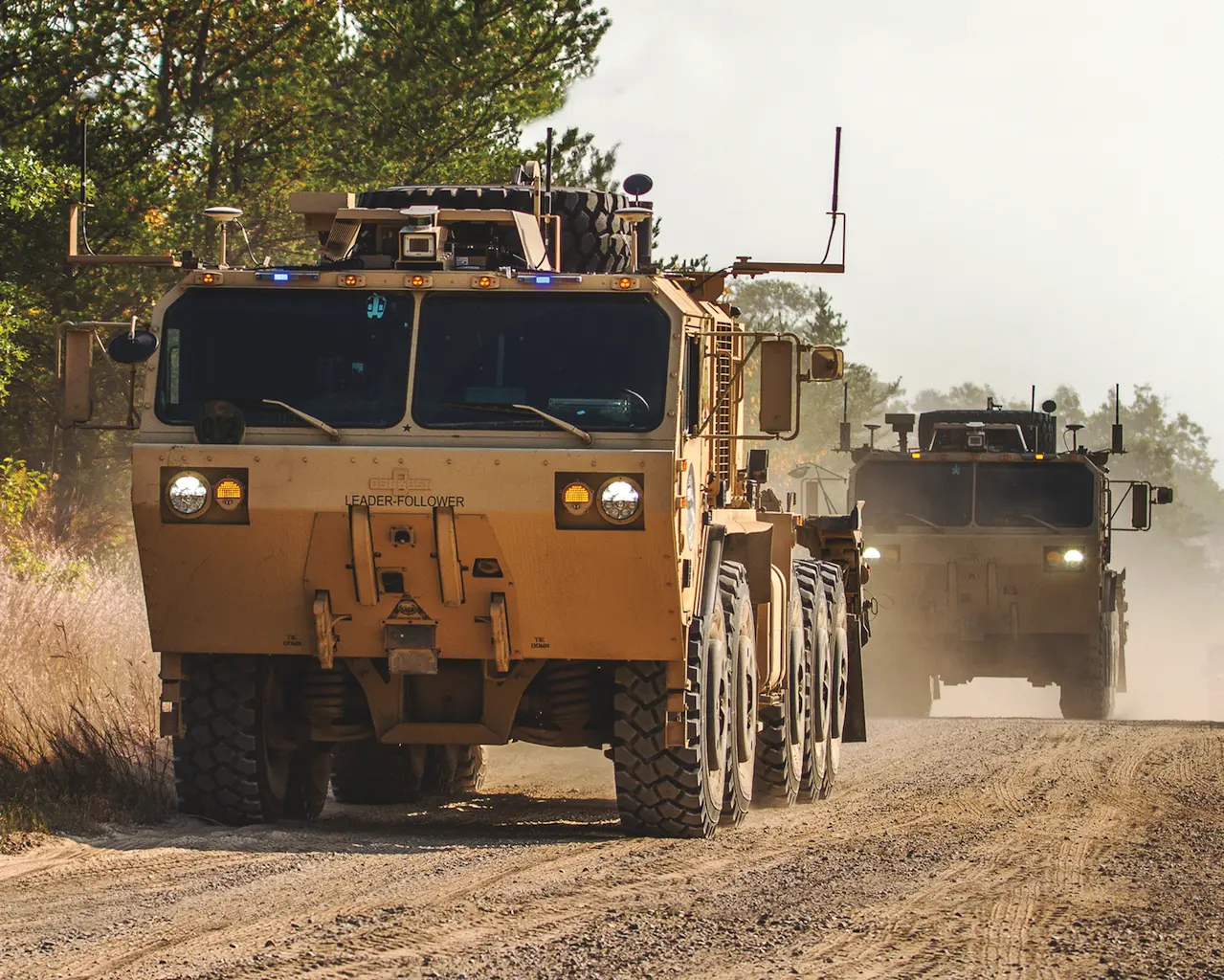 Two Oshkosh PLS A1 heavy trucks driving in convoy on a dirt road